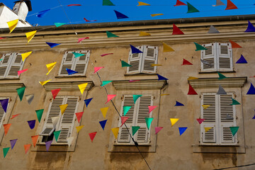 Multicolored pennant flags strung across a beige building with white window shutters under a deep blue sky, creating a festive street atmosphere.