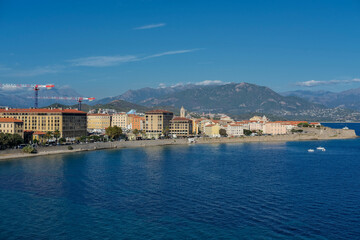 Coastal cityscape with colorful buildings and mountains in the background, viewed from the sea. Boats and calm blue water under a clear sky.