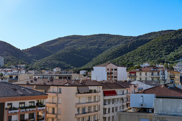 Urban rooftops with red tiles and green hills in the background under a clear blue sky. Mediterranean town with residential buildings and mountain scenery.