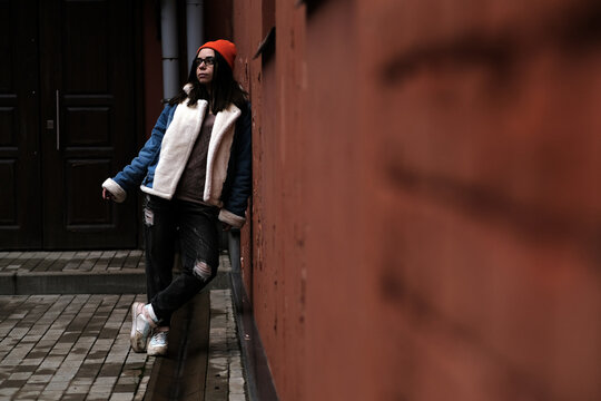 A young woman leans against a wall in a stylish outfit featuring a cozy jacket and vibrant beanie She enjoys a moment of relaxation on a brisk day in an urban area