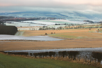 A patchwork of frosty fields in rural countryside stretches across the rolling farmland near Kennoway in Fife, Scotland with a view to the distant Lomond Hills on a calm winter morning.
