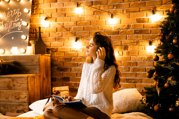 beautiful curly girl in a white sweater with dark hair sits on the bed smiling and writes in a...