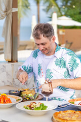 Man enjoying tropical dining outdoors near beach, wearing floral shirt, surrounded by diverse dishes and chocolate drink. Leisure, vacation, culinary, beachside, relaxation.