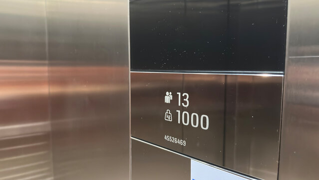 Elevator capacity sign showing maximum 13 persons and 1000 kg load on stainless steel panel, modern lift safety display in hotel, apartment, office, mall or commercial building interior