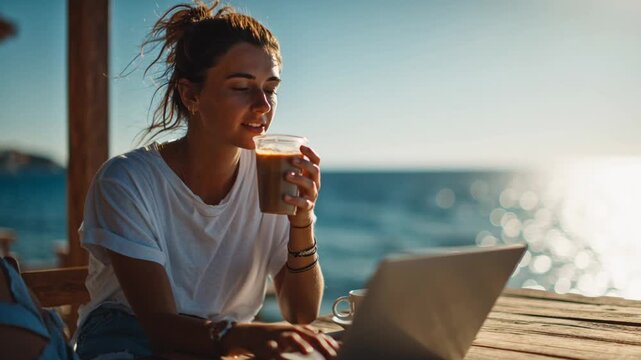 Young woman working on laptop and drinking iced coffee by the sea