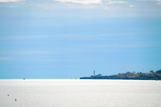 A distant lighthouse stands along a rocky coastline beneath soft blue skies and reflected sunlight, symbolizing guidance and calm maritime simplicity.