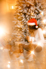 blurred New Year's background. beautiful curly girl in a white sweater with dark hair sits on the floor and holds a gift on the background of a Christmas tree