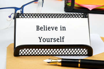 On a desk, motivational words of encouragement reading Believe in Yourself are presented, beside a pen and framed by eyeglasses and sticky notes