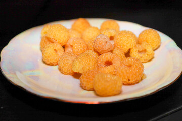 A cluster of ripe yellow raspberries is vividly displayed on a light, iridescent plate, creating an appetizing close-up shot. The warm orange hues of the berries contrast with the dark background and 