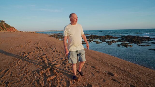 Hombre mayor con problemas de movilidad camina y sonr&iacute;e por la playa al atardecer.