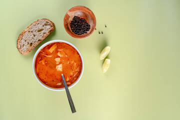 A top table view of bowl of soup with a spoon in it on a green table. The bowl is filled with a red. A slice of bread and pieces of lime are also present on the table