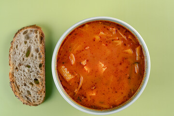 A bowl of soup sits next to a piece of bread. The soup is red and he is a hearty meal. The bread is to the left of the soup