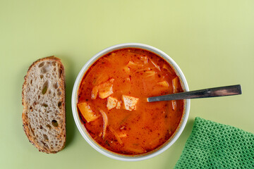 A close up of bowl of soup sits on a green table next to a piece of sourdough bread. The soup is red and has a spoon in it. Tofu and some vegetables are visible in the soup. Green cloth on the side