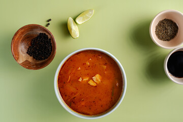 A bowl of soup sits on a table with a few other bowls and a spoon. The soup is a bright orange color and is surrounded by spices and other ingredients. Concept of warmth and comfort