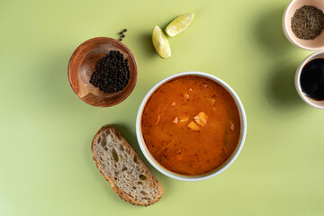 A bowl of soup sits on a green table with a loaf of bread and a few bowls of spices