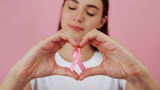 Young Woman's Hands Form Heart Around Pink Ribbon for Breast Cancer Awareness and Support on Pink Background