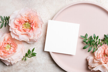 Square blank wedding card mockup on a round beige plate with peach roses and green leaves on a textured background. Elegant flat lay for white invitation or greeting cards