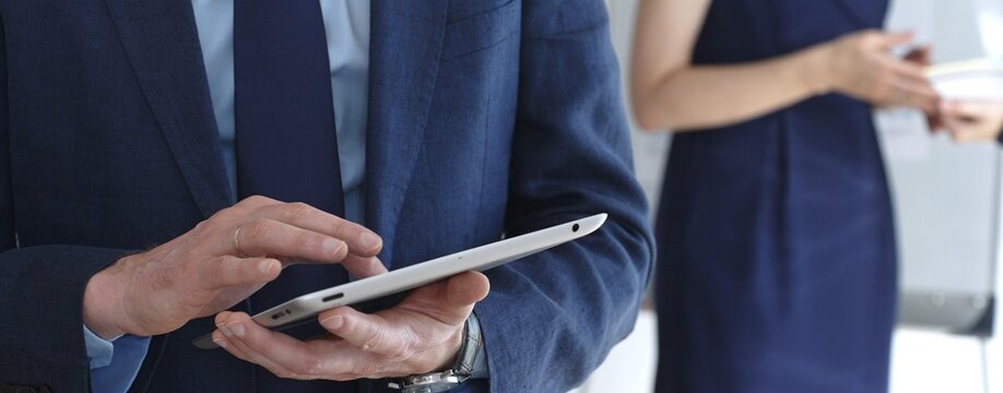 Businessman in a blue suit using a tablet during a corporate meeting, interacting with the touchscreen as blurred colleagues collaborate in the background, in office. Business people concept