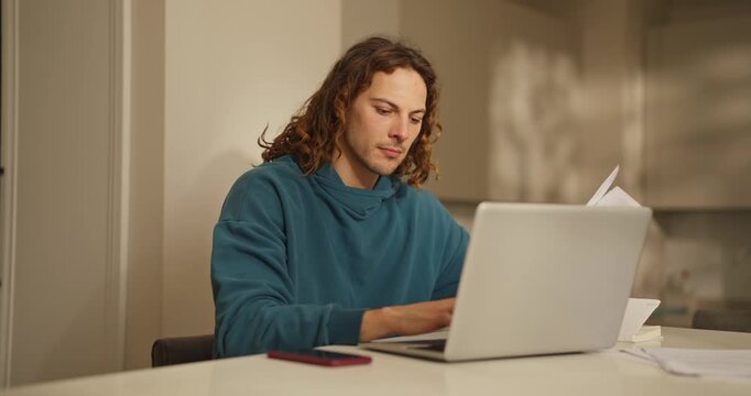 Young man in a hoodie sits at a table, concentrating on his laptop while organizing scattered papers in a bright room