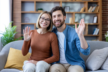 Happy diverse couple sitting on a sofa at home, smiling and waving hands at the camera during an online video call, symbolizing remote connection and communication