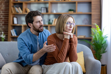 Man reaches out to upset partner on sofa as she turns away, tense couple after disagreement in living room, emotional moment of apology, support and attempted reconciliation