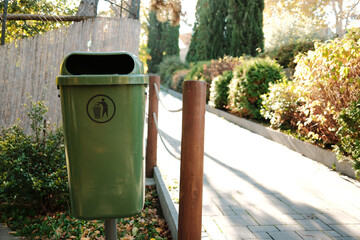Green trash bin standing beside sunny pathway with plants and trees. Clean environment and eco-friendly concept