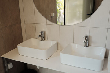 Two modern white sinks with chrome faucets and large round mirror inside tiled restroom
