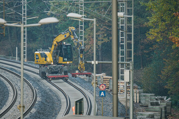 Excavator with an arm on railway tracks, a train in the distance between electric poles.
