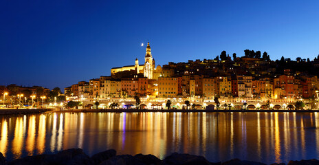 Panoramic night view of Menton, France, with illuminated old town reflecting on the water, and the...