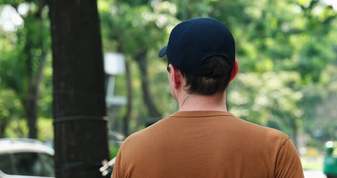 Rear view man wearing cap walking in park, casual outdoor setting, summer or spring day, greenery background, peaceful ambiance.