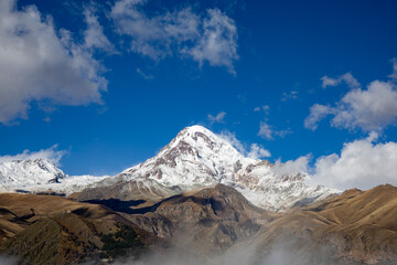 A breathtaking view of the Mount Kazbegi, featuring its snow-covered summit sharply contrasted against a deep blue sky and surrounded by dry, rocky mountain slopes and patches of low-hanging mist