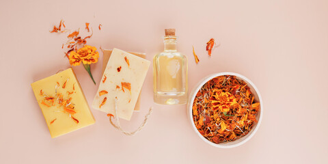 Flatlay of handmade soap bars with dried calendula petals, marigold flower, herbal oil bottle and ceramic bowl on pastel background
