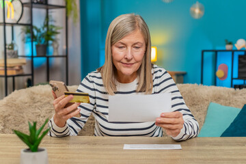 Mature woman uses credit card and smartphone to transfer money pay utility domestic bills invoices at home. Grandmother manages financial documents, handling transactions with focus and determination