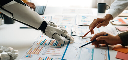 A robot arm reaches out as a human collaborates on data-driven graphs and charts in a modern office setting, symbolizing teamwork and technological integration in business decisions.Noogenesis