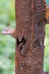 Vertical close-up of a heavily corroded and pitted brown metal post featuring a visible hole and sharp edges, backed by a soft, blurred green background