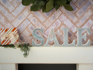 Brick fireplace hearth mantle Christmas holiday festive decoration with Sale letters and wrapped gift, hanging green wreath and white wood shelf.