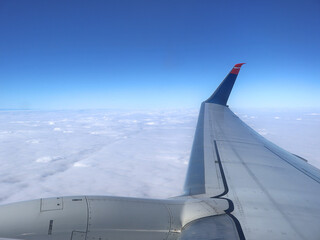 A passenger jet wing slices through clear blue air above a sea of clouds, offering a serene, high-altitude perspective that evokes travel, freedom, and the adventure of flight.