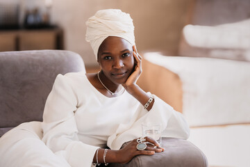 A pensive woman in a white turban and dress lounges on a sofa, holding a glass, embodying elegance and thoughtful reflection.