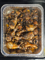 Several seasoned chicken drumsticks and roasted mushrooms are presented in a silver foil pan, ready to be served.