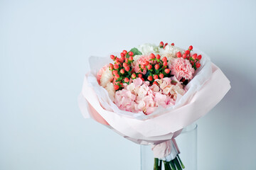 Elegant bouquet of white hydrangeas, pink carnations, and red berries wrapped in pastel pink paper, placed in a glass vase
