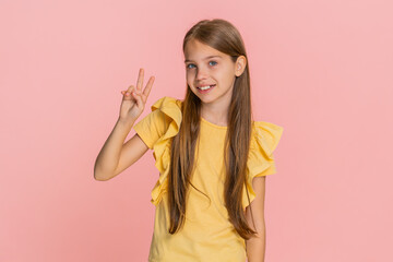 Young child girl makes a peace sign with both fingers while smiling warmly at the camera, radiating joy and positivity. School kid isolated on pink background, cheerful and full of youthful spirit.
