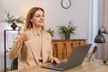 Middle-aged businesswoman at home office checks results on laptop smiles shows thumbs up gesture...