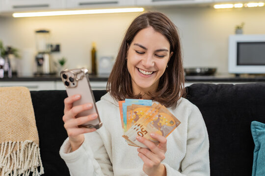 Young woman at home counts euro cash checks rate in phone app smiles and plans big buy goal aim. Caucasian girl on sofa happy about salary and bonus plans invest saves money and notes amount in budget