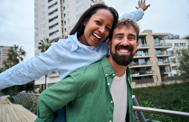 Cheerful bearded man giving a joyful piggyback ride to a smiling woman in a vibrant city setting, celebrating their love and happiness while enjoying a playful moment together looking at camera