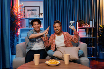 Couple gamer making high five holding joysticks be winner together complete level sitting sofa playing video game with front snack and drinks at neon light color living room at modern home. Infobahn.
