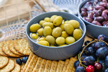 bowl of green green and calamata olives, cheese, served with crackers and other fruits The board is filled with various snacks, including crackers, grapes,