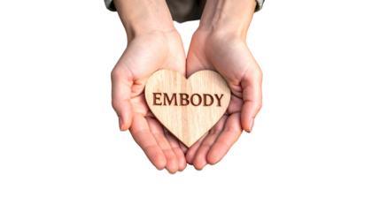 Person cupped hands holding a wooden heart with "EMBODY" etched. Black background
