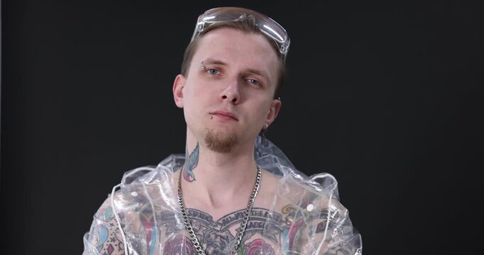 Young man with piercings, tattoos and stylish clothes posing on dark grey background. Camera moving backwards