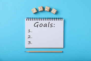 New 2026 Year Goals. Inscription in notebook and cubes with numbers on light blue background, top view