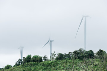 Giant wind turbines generate green electricity in a mountainous landscape. Under a cloudy and foggy sky, the turbines can be seen penetrating the clouds.